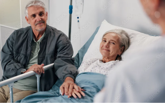 Patientin liegt im Krankenhausbett und lächelt, während ein Angehöriger ihre Hand hält und dem medizinischen Personal zuhört.
